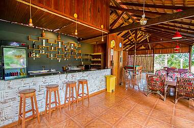 Bar der Turtle Lodge mit hölzernen Barhockern an der Theke, einem Kühlschrank und einem Weinregal hinter der Theke und Holzmöbeln und einem Billardtisch im hinteren Bereich 